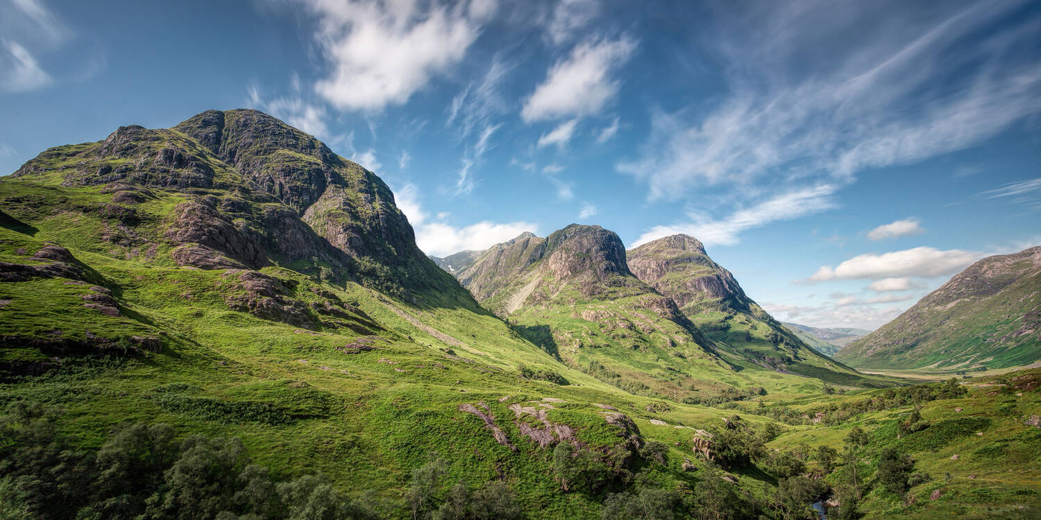 A view looking down Glencoe in summer on a sunny day. The mountains are green, and white clouds streak across the sky.