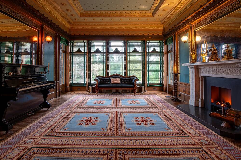 A view of an extremely ornately decorated drawing room, with the far wall made up of floor-to-ceiling windows. A red and blue patterned carpet almost fills the floor on top of parquet wooden floorboards. The ceiling has decorative, gilded plasterwork. There is a piano to the far left of the room, and a fireplace on the right-hand wall. Large mirrors run either side of the room.