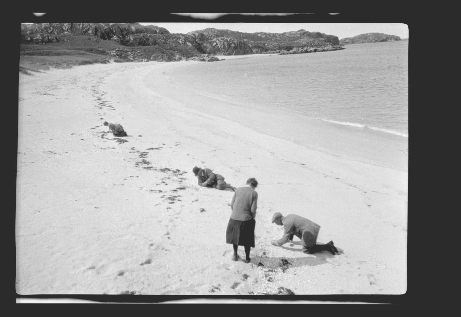 A black and white photo of four people looking for shells on a sandy beach. Three sit or crouch on the beach, running their hands through the sand. The fourth woman stands and leans over the man closest to the foreground.