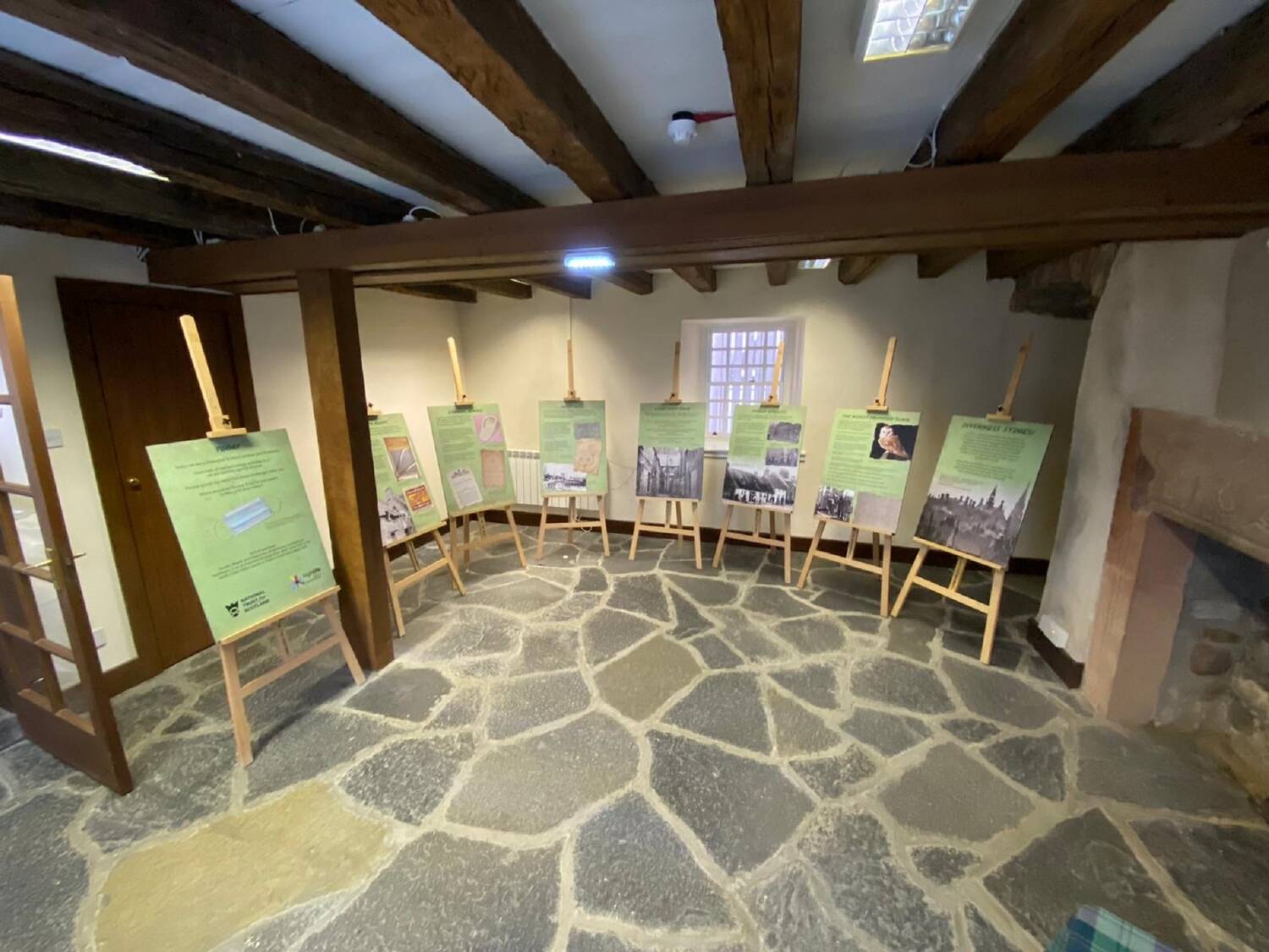 A room in an old house with a flagstone floor and exposed timber beams in the ceiling has an exhibition on display. The exhibition is shown on various panels mounted on wooden easels around the edge of the room.
