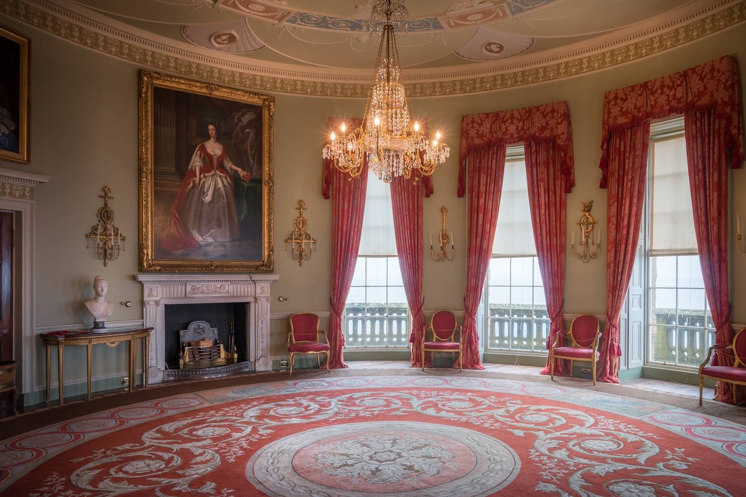 A view of the Round Drawing Room at Culzean Castle, showing the circular room. Floor-to-ceiling red curtains hang at the three large windows. Further round large, gilt-framed portraits hang on the wall above the fireplace. A large circular red patterned rug fills the floor.