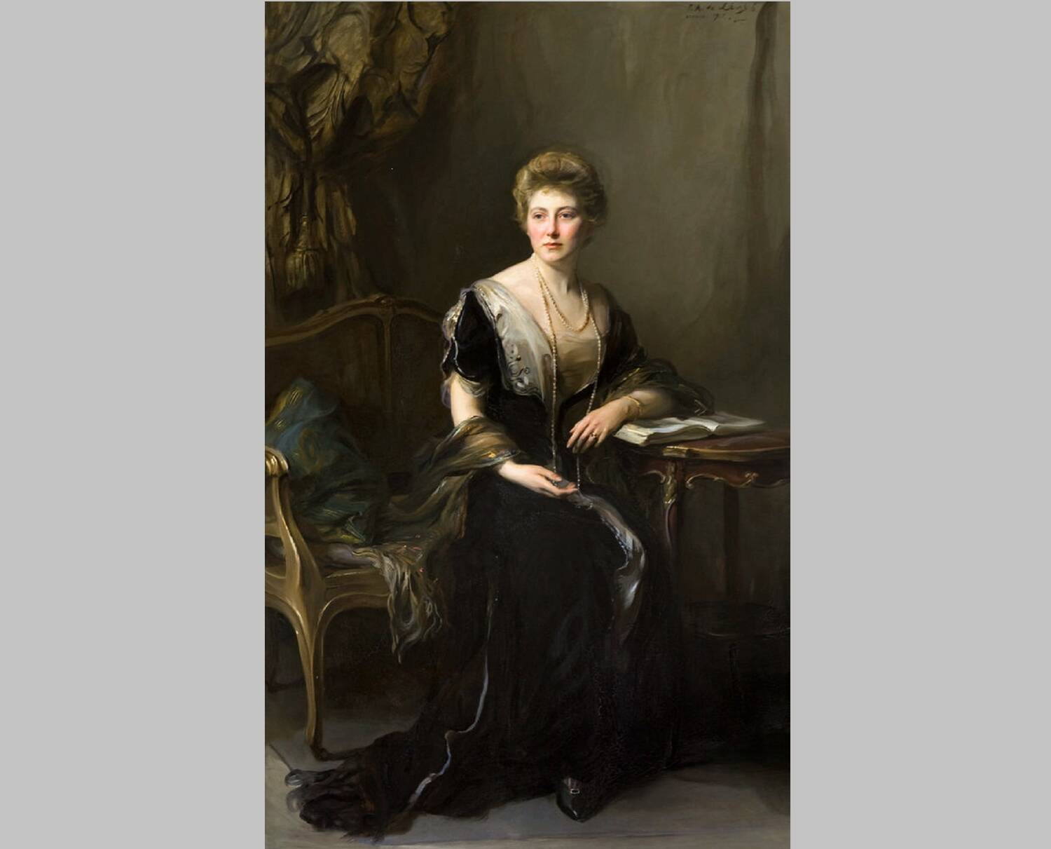 A oil painting of a young woman, dressed in a long dark dress. She is seated at a small table, and rests her arm on an open book on the table. Her hair is arranged in top of her head, and she wears a long pearl necklace.