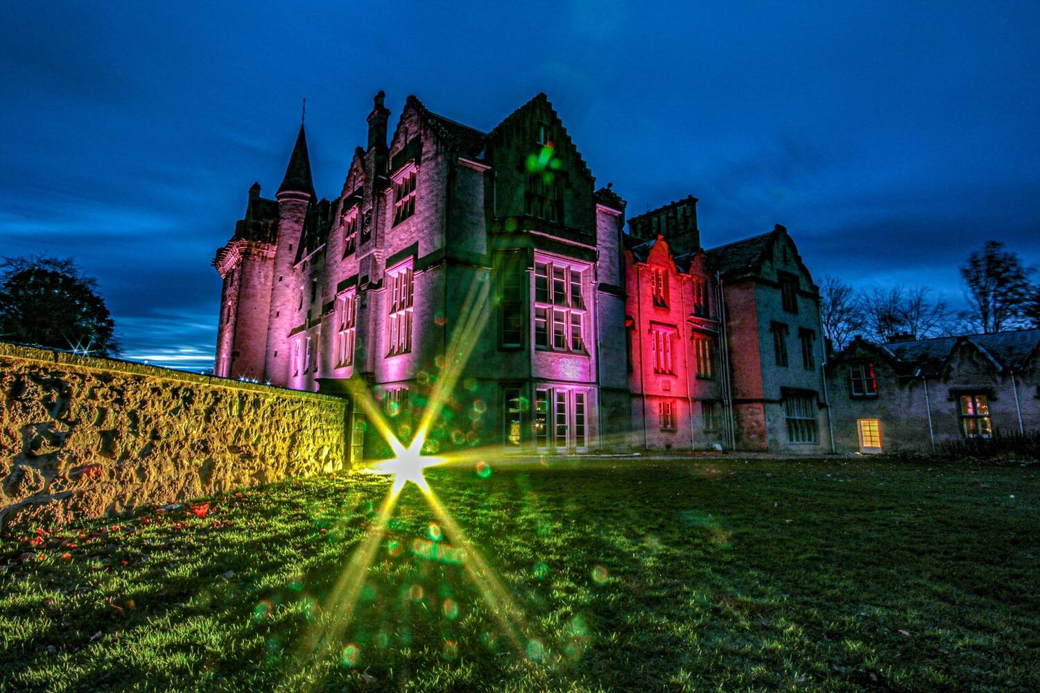 A castle is lit up with multi-coloured lights at night time. A long stone wall and lawn area feature in the foreground.