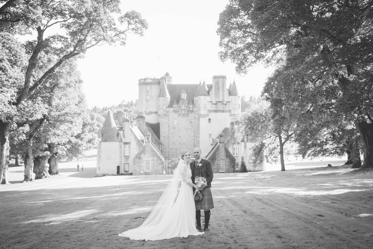 A black and white photograph of a bride and groom standing on a lawn, with Castle Fraser in the background. Tall woodland trees frame the shot. The bride wears a veil and her long white train is arranged around her. The groom wears a kilt. Both hold a bouquet of flowers between them.