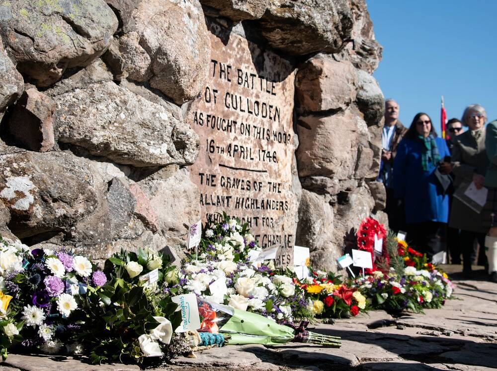 Wreaths and bunches of flowers are displayed at the base of a large memorial stone. The stone has the following text inscribed on it: The Battle of Culloden as fought on this moor 16th April 1746.  A group of people can just be seen standing to the side of the stone.