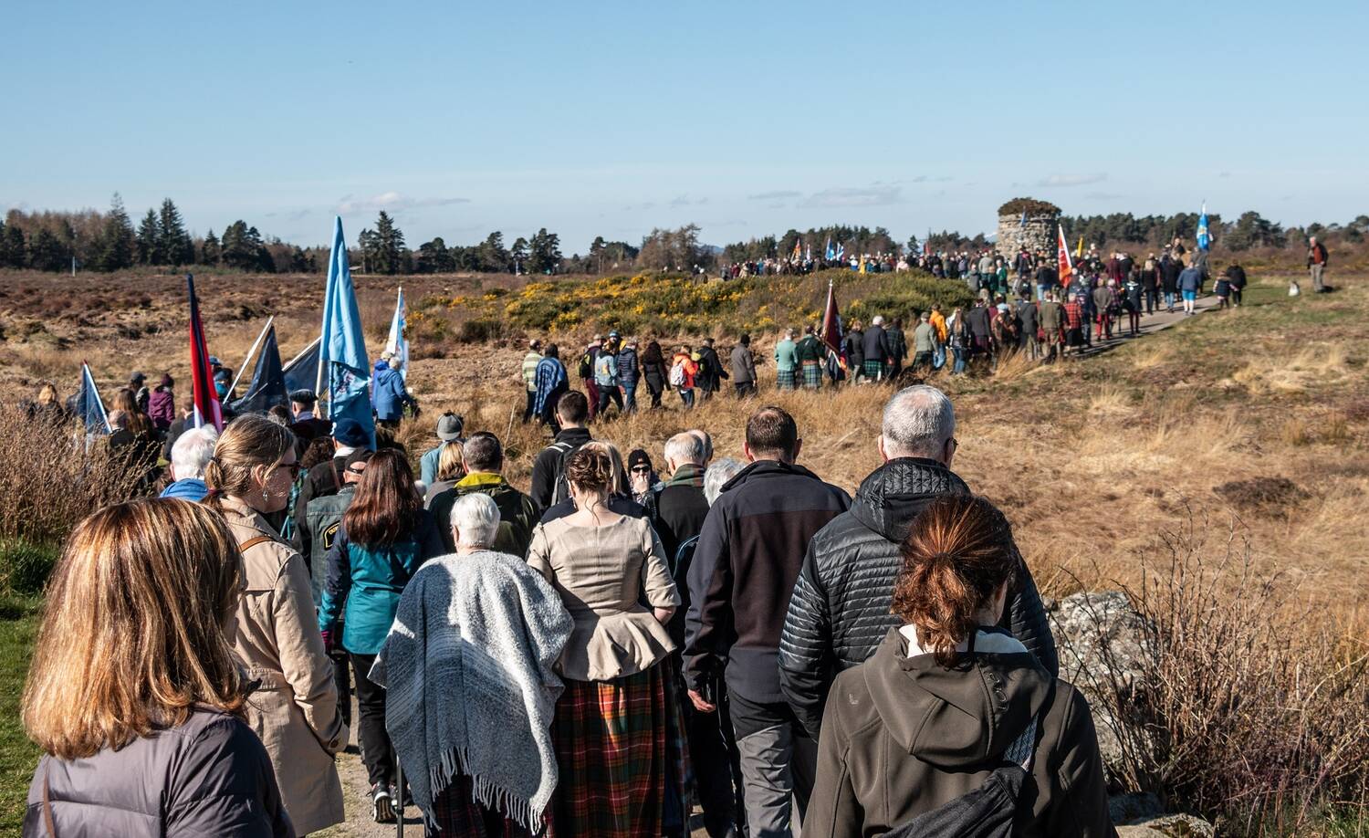 A long line of people snake along a footpath across Culloden Moor towards a large stone cairn in the distance. Many are carrying flags.