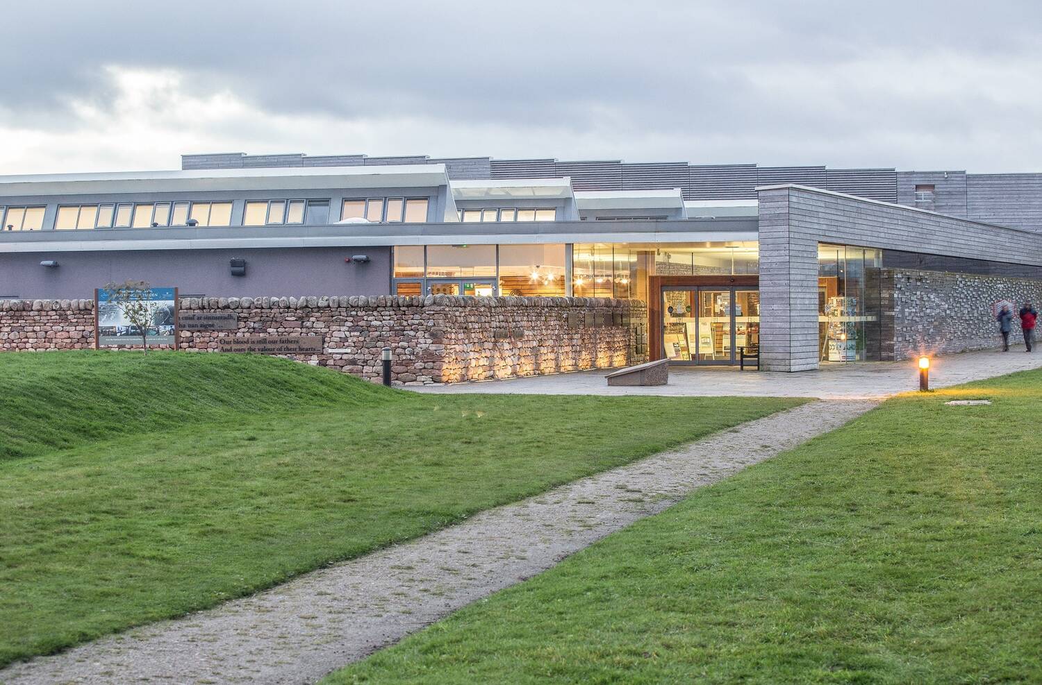 A view of Culloden visitor centre, looking towards the entrance from a path leading across a grassy area. The centre is lit inside with a welcoming orange glow.