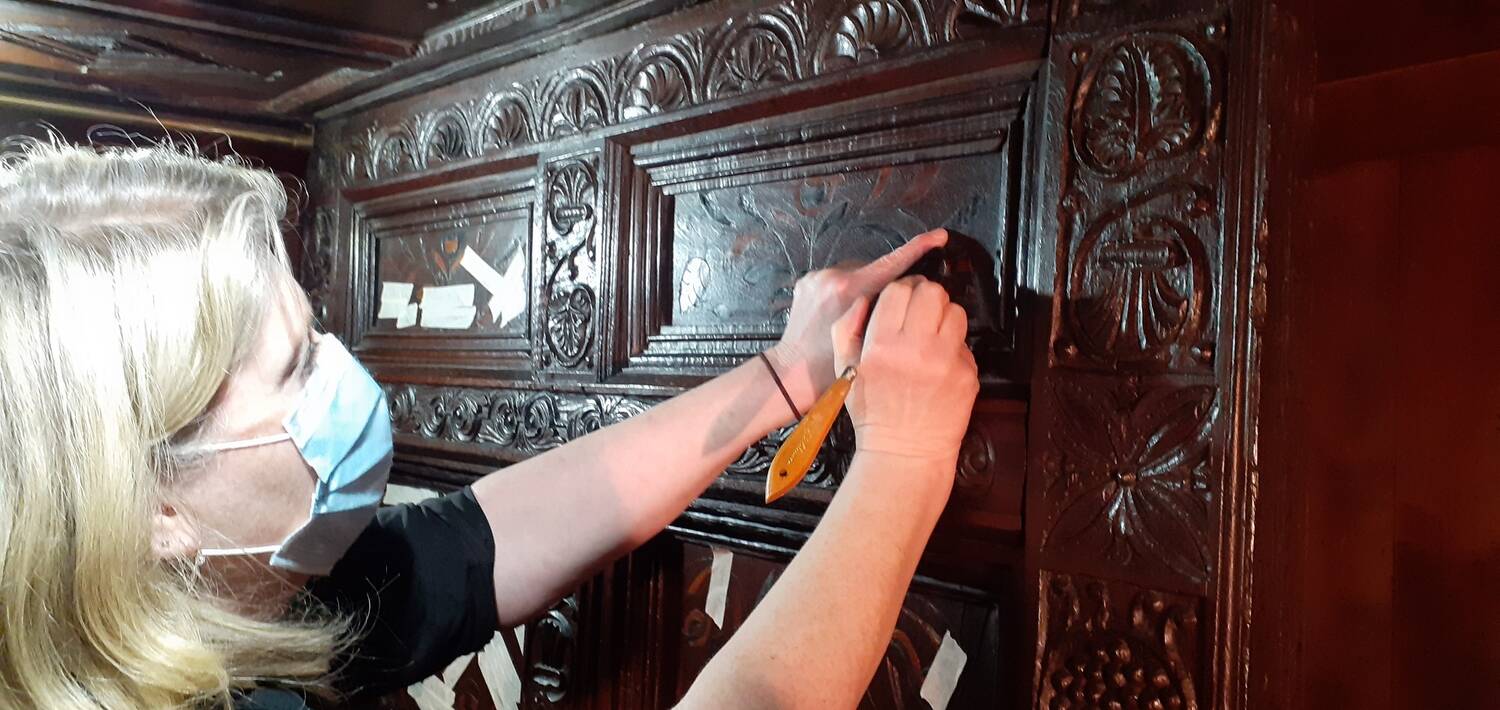 A lady wearing a face covering uses a small tool to make repairs to a carved wooden headboard of a bed.