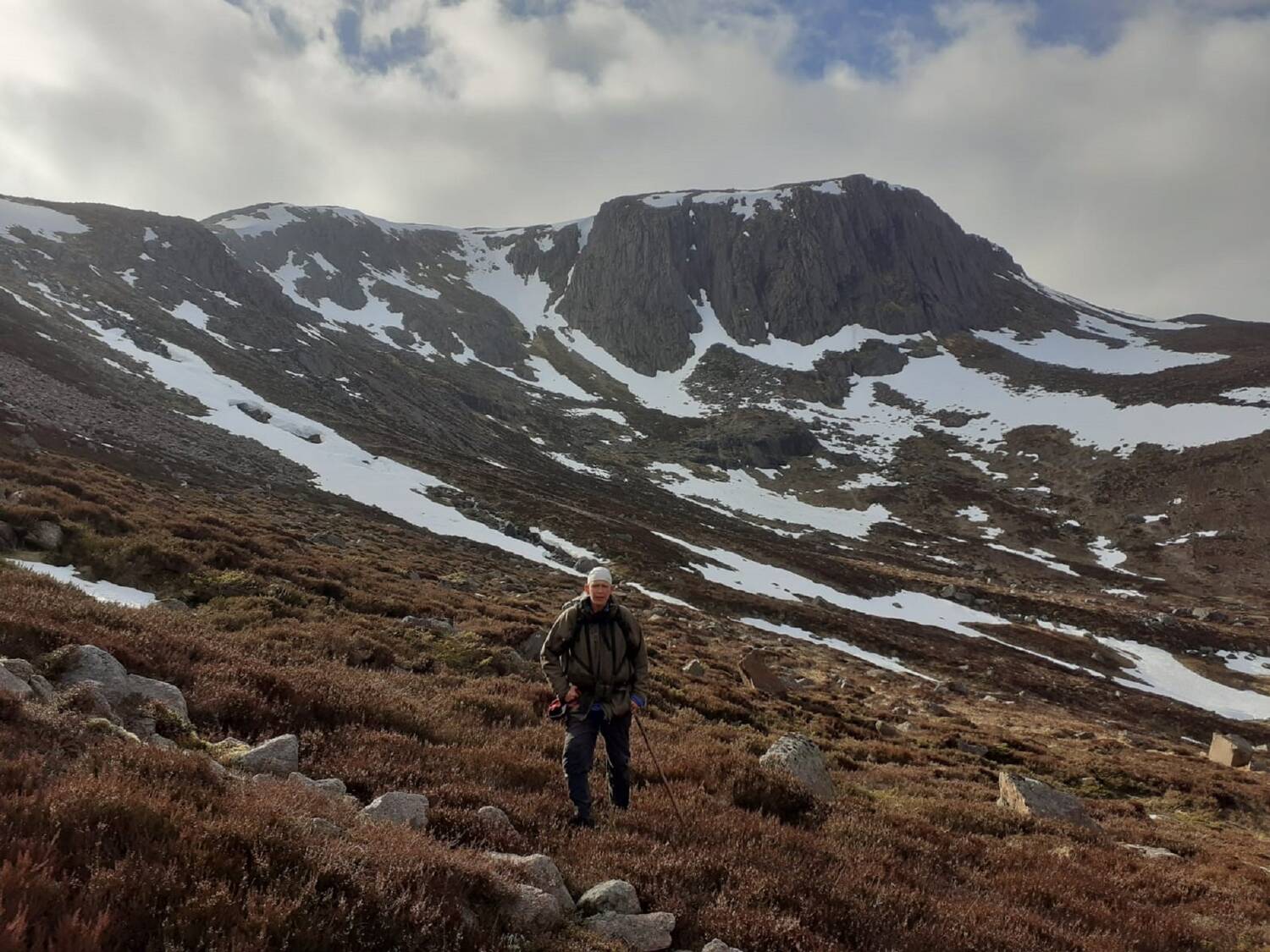 A woman stands on a heather-covered hillside strewn with boulders. Behind her is a deep wide glen and steep, snow-pocketed mountains. The sky is heavy.