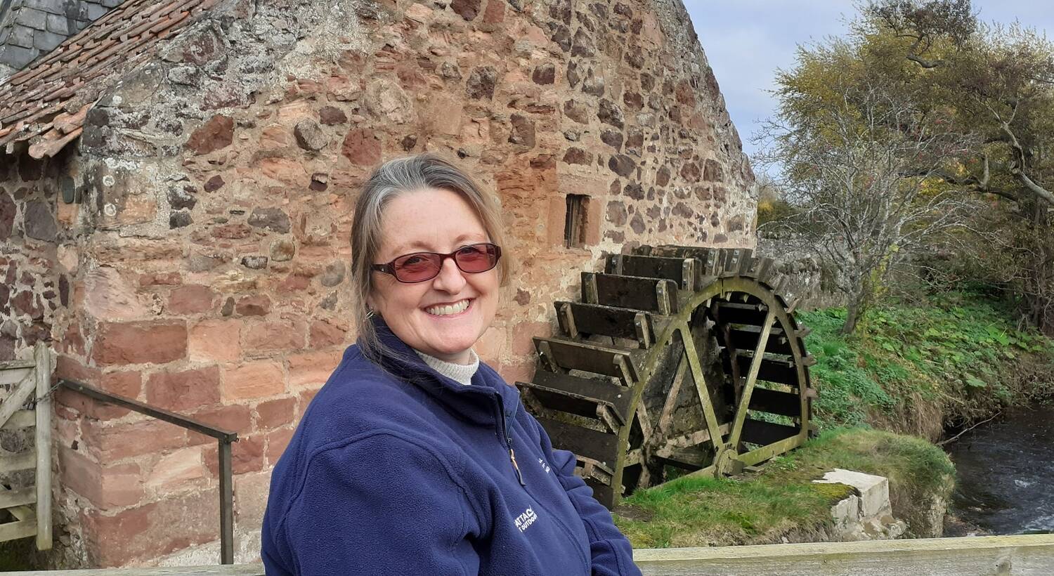 A smiling woman wearing a navy fleece jacket stands beside a water wheel at the side of an old mill building.