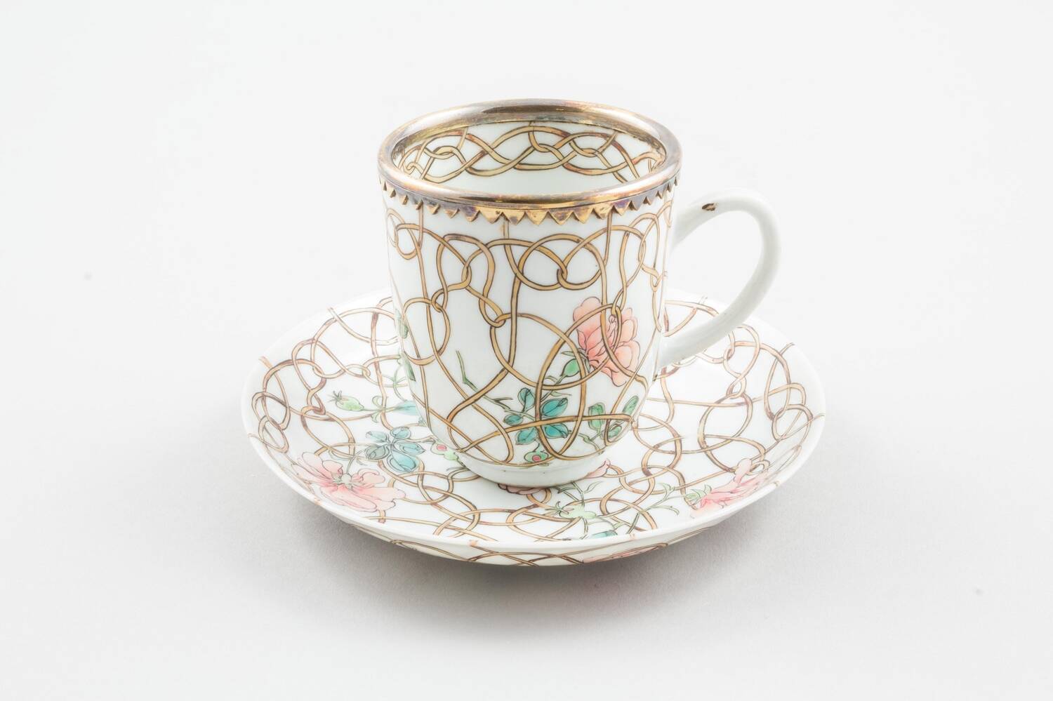 A tea cup sitting on a matching saucer is displayed against a plain grey background. It has a silver-gilt mount and is decorated with pink flowers and gold tendrils. 