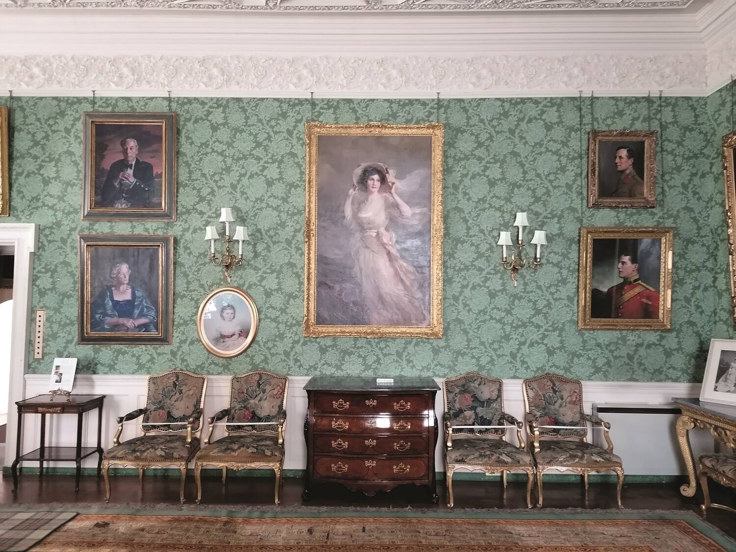 A portrait of a young Edwardian woman, wearing a floaty white dress and a large bonnet, hangs at the centre of a collection of paintings on a green-papered wall. Smart chairs and a polished wooden chest stand below the group of portraits.
