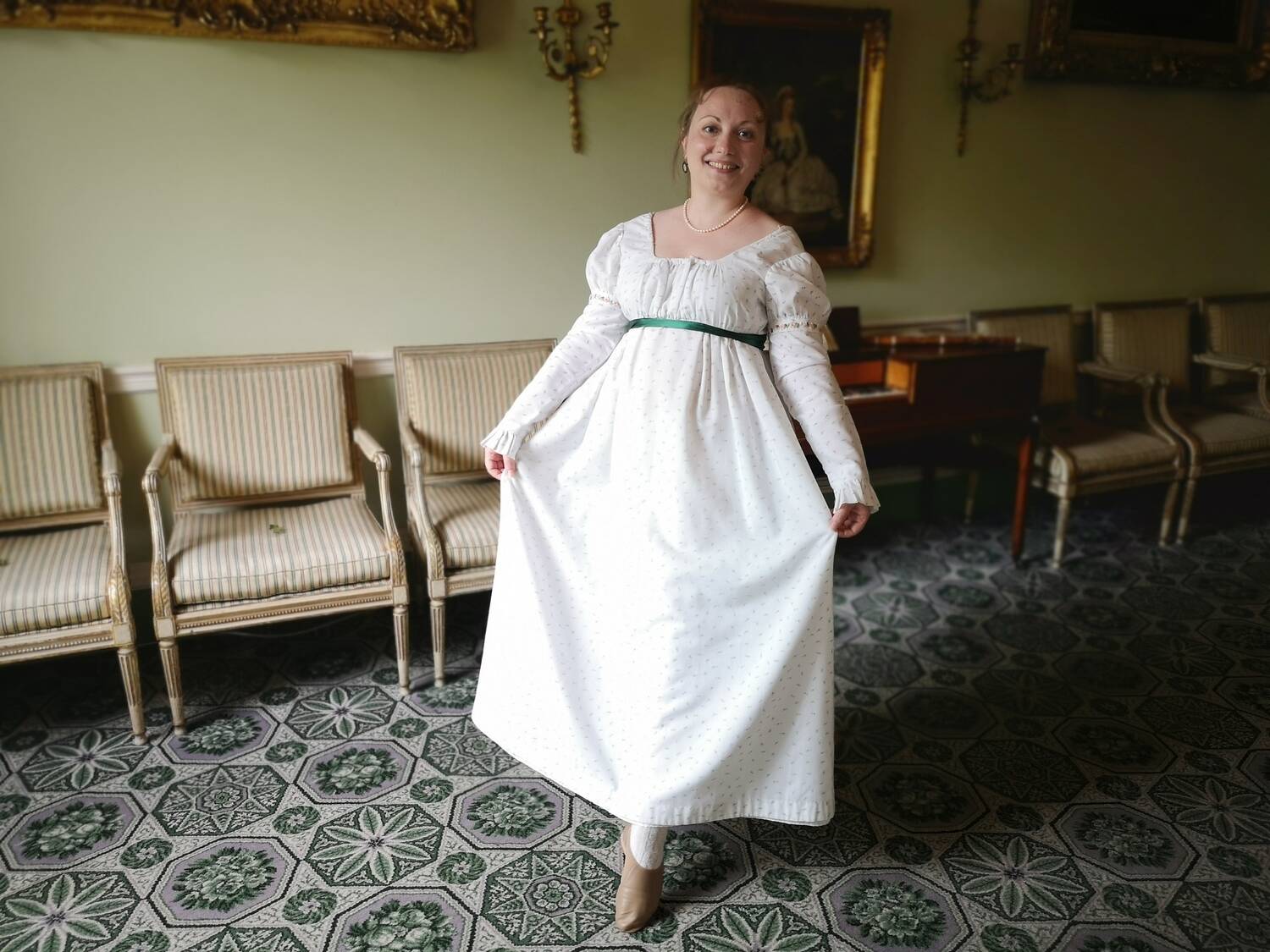 A woman in a white 18th-century dress with a green ribbon under the bust holds her skirts and points her foot forward and to the floor, as if about to dance.