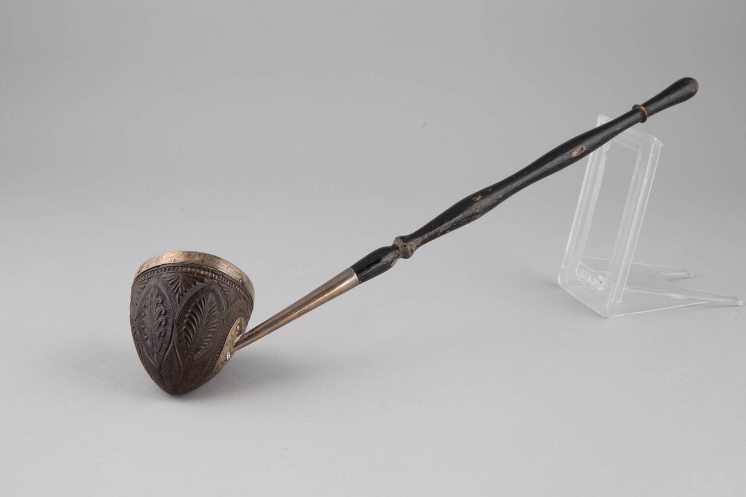 A small, silver-mounted coconut shell ladle with a black painted wooden handle is displayed against a plain grey background, resting its handle on a perspex holder.