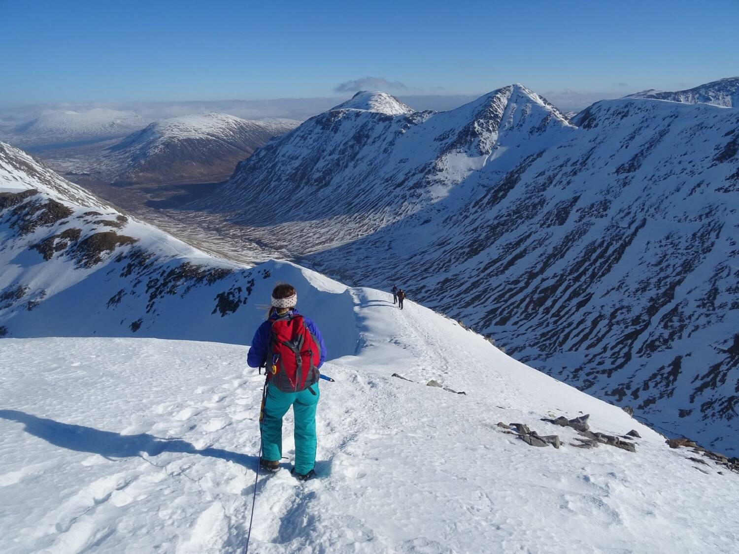 A woman dressed in full winter gear stands almost at the summit of a snow-covered mountain, looking down towards a ridge path, with the glen far below. The sky is bright blue and the snow is glistening.