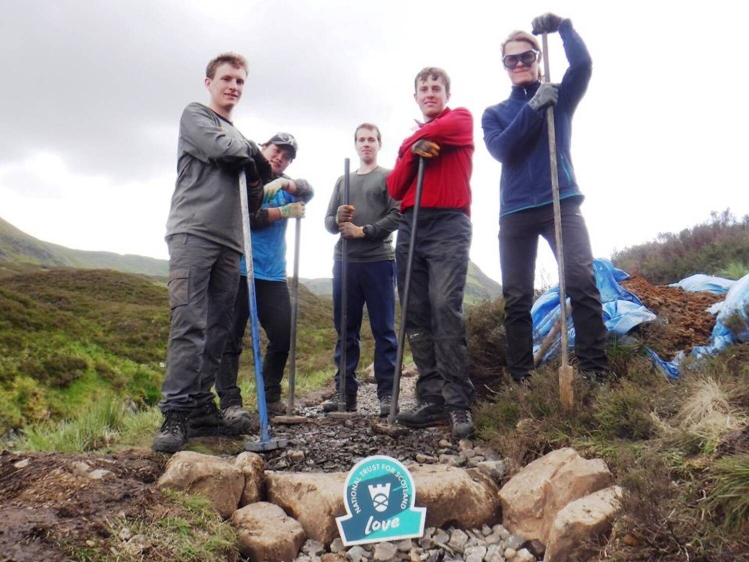 Five people stand beside a mountain path, holding long tools used to shape the path. A National Trust for Scotland ‘Love’ sign sits in the foreground, on the path.