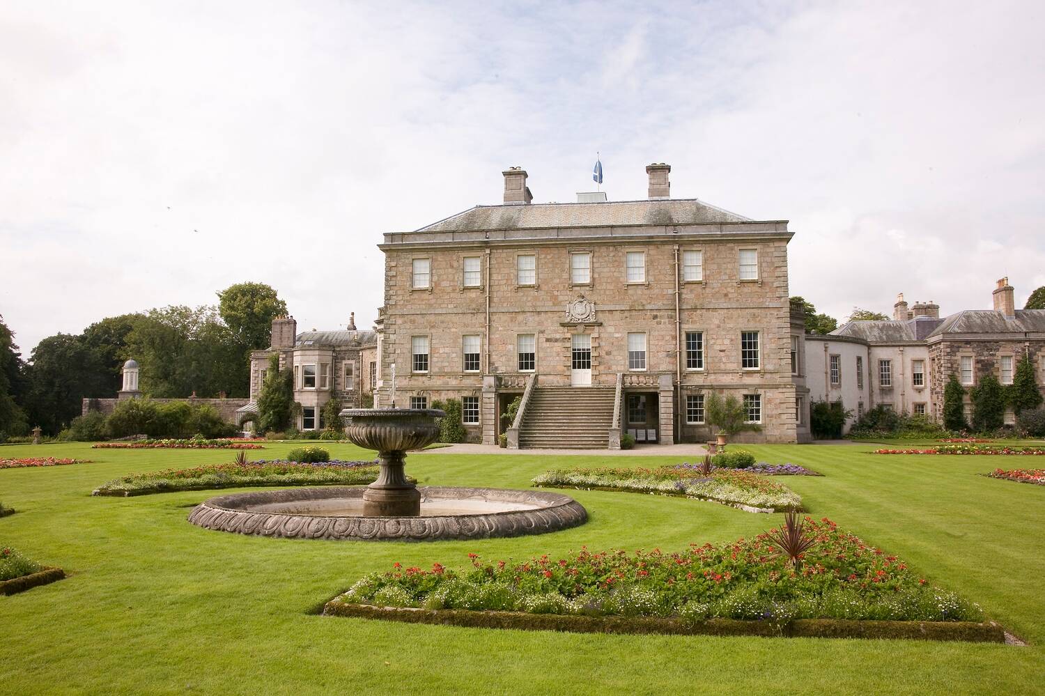 A view of a large Georgian country house with immaculate lawns in front. At the centre of the lawns and flowerbeds is a circular fountain, gently trickling.