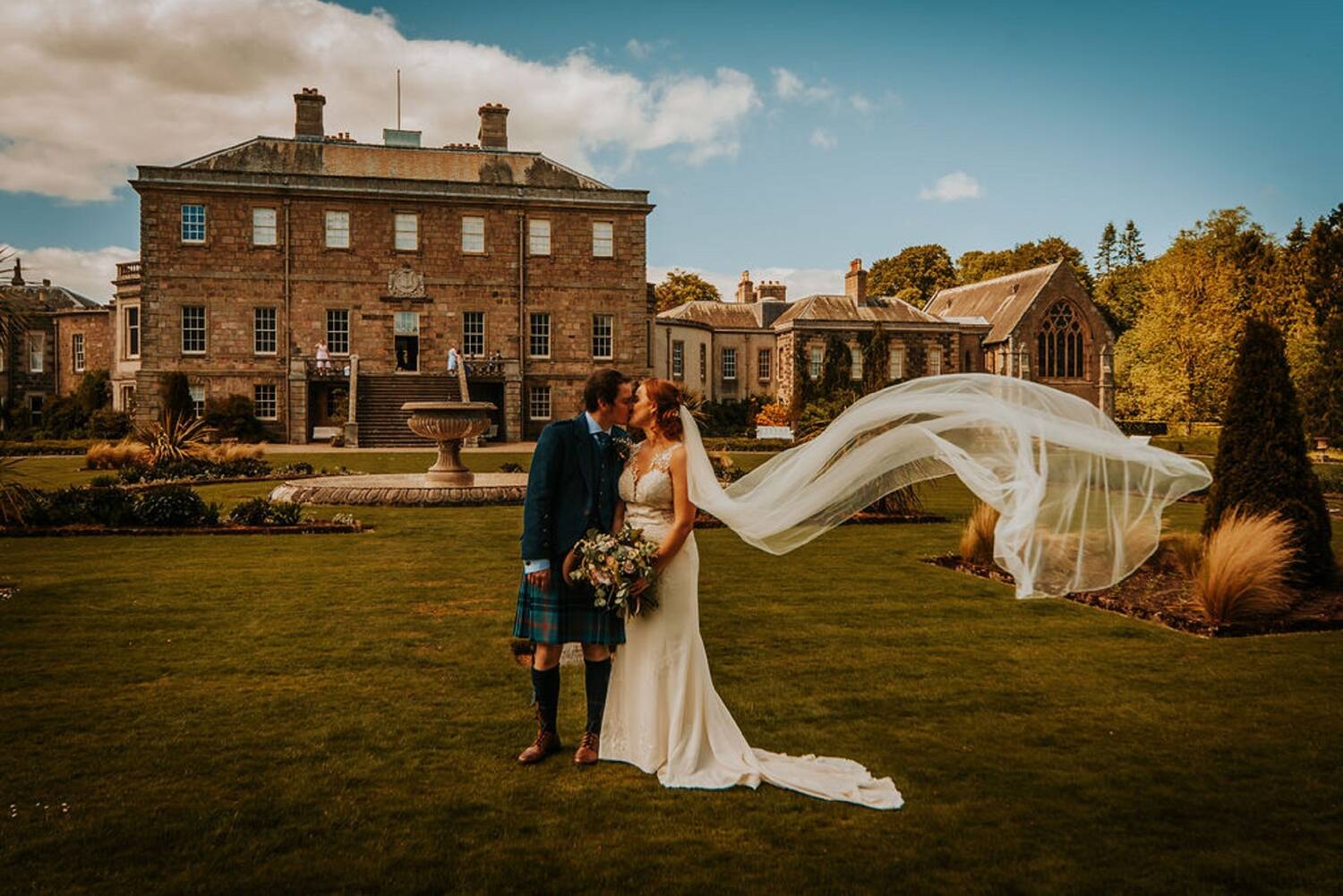 A bride and groom stand kissing on a lawn in front of a grand stately home. A fountain is just behind them. The wind is catching the bride's long veil, blowing it away from her.