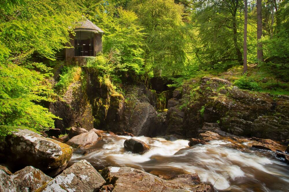 A little stone folly stands on rocks on top of a rushing waterfall. It is surrounded by bright green leafy trees. The river rushes over large rocks and boulders in the foreground.