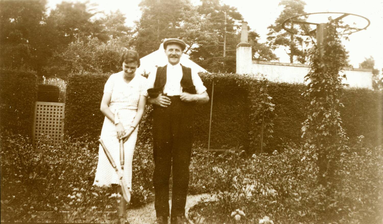 A black and white photo of a man and a woman standing side by side on a narrow path in a rose garden. The woman is on the left - she wears a white dress, holds a very large pair of shears and is looking down at her feet. The man is on the right - he smiles directly at the camera and is holding his dark waistcoat over his white shirt. He also wears a flat cap.