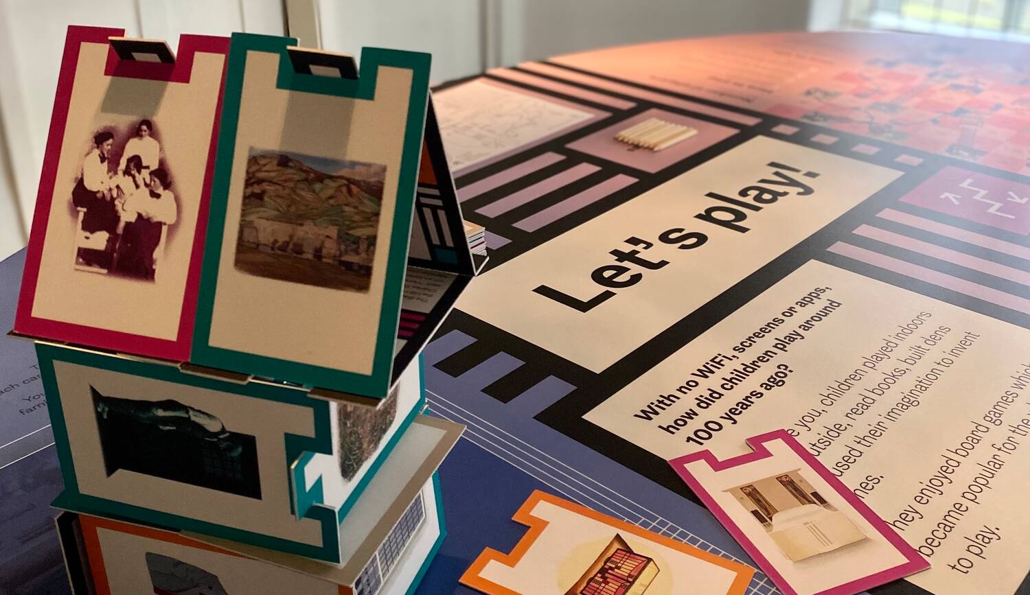 A piece of a new immersive installation, with a colourful display board reading 'Let's play!' and a house constructed out of large individual cards, each one decorated with a different picture relating to the Hill House and its history.