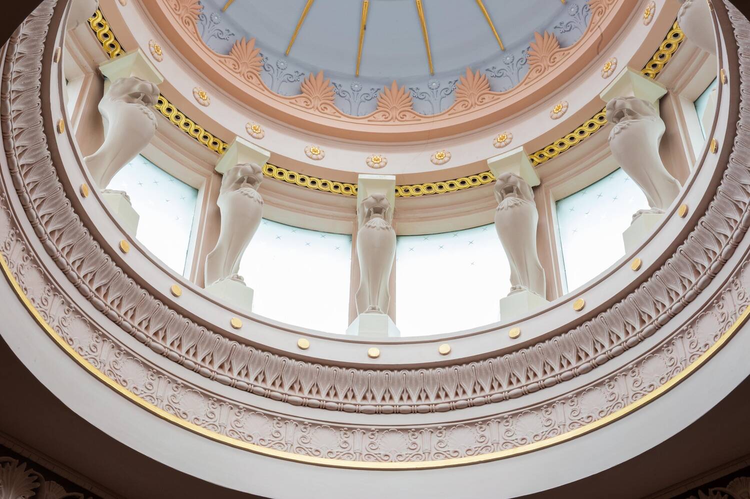 A view of the inside of a painted dome, with blue and gold decoration at the very top. Statues separate small window panels all the way around.