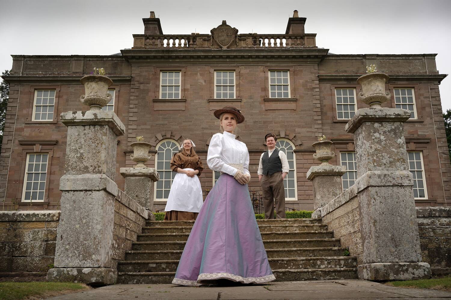 Three costumed guides dressed in various Victorian outfits​ stand on the stone steps leading up to a grand Georgian mansion house. There is a cook on the left, a well-to-do young woman in the centre and an estate worker on the right. Stone urns with pretty bedding plants line the staircase.