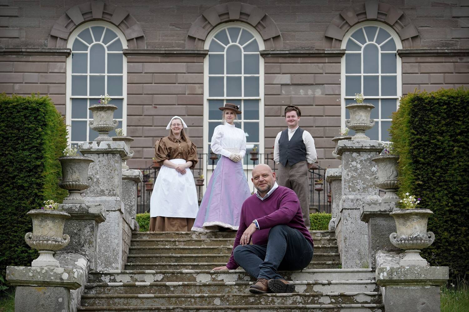 A man sits on the stone steps leading up to a grand Georgian mansion house. Behind him stand three people, dressed in Victorian costumes: a cook on the left, a well-to-do young woman in the centre and an estate worker on the right. Stone urns with pretty bedding plants line the staircase.