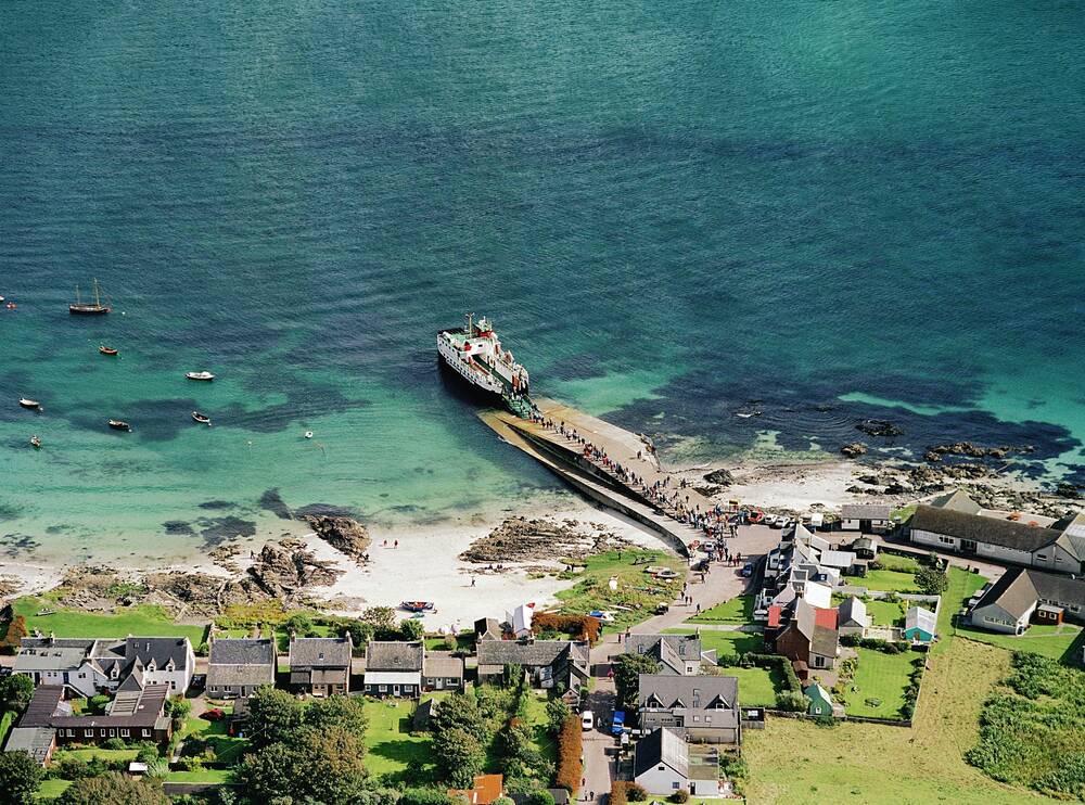 An aerial view of the pier on Iona, showing a ferry docked. A row of houses can be seen along the shoreline. The sea is turquoise and clear.