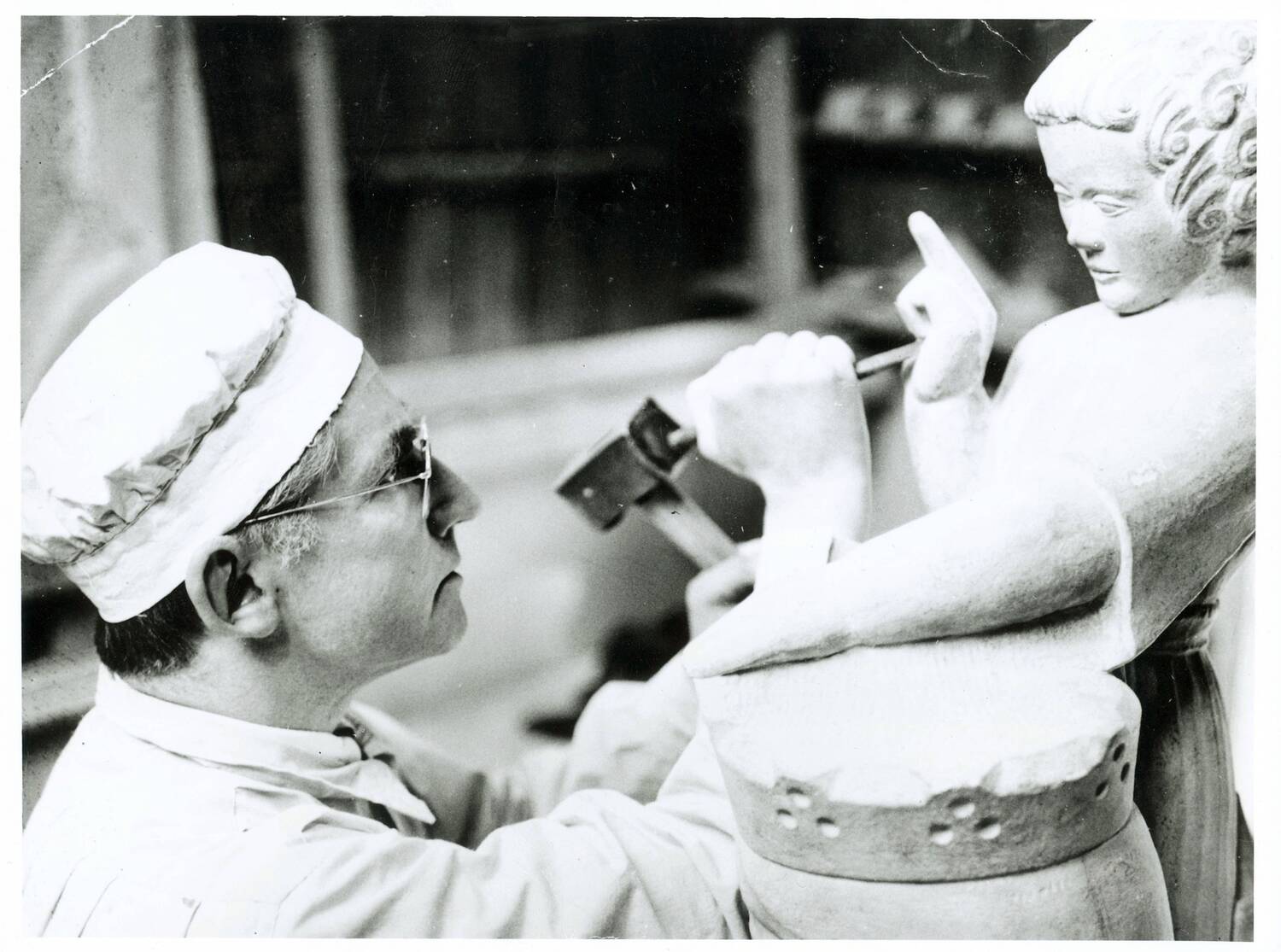 A black and white photo of a young man working on a sculpture of a cherub-type figure. He holds a small stone chisel and is working on the figure's hand. The statue and the man appear to be looking at each other eye to eye.