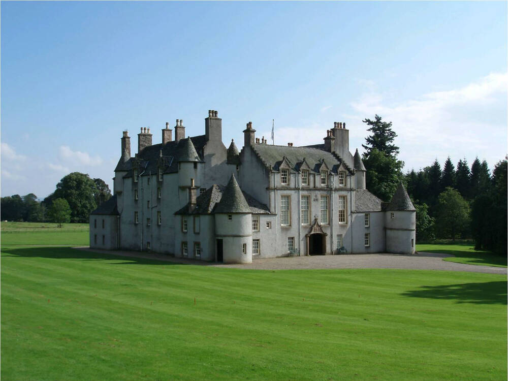 A view of the exterior of Leith Hall, from across some manicured lawns. It is a large stately home, with grey walls and a slate roof, with little towers and turrets at each corner.