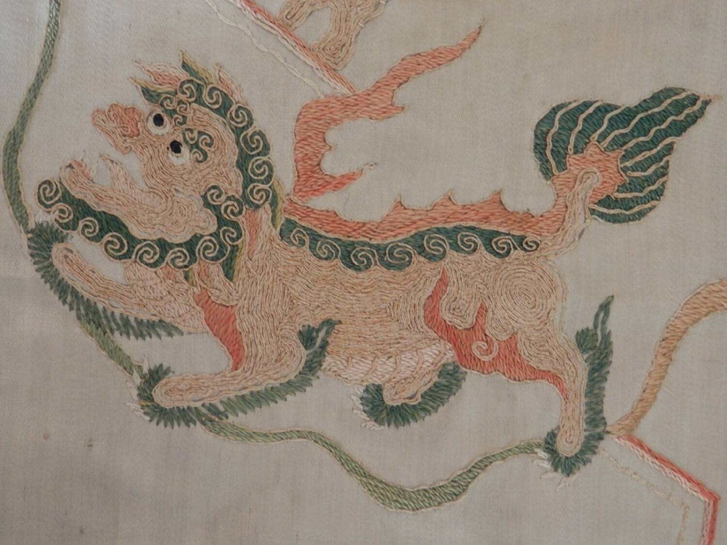 A close-up image of an embroidered Chinese-style lion on a silk panel. The lion is sewn in green, red and orange thread.