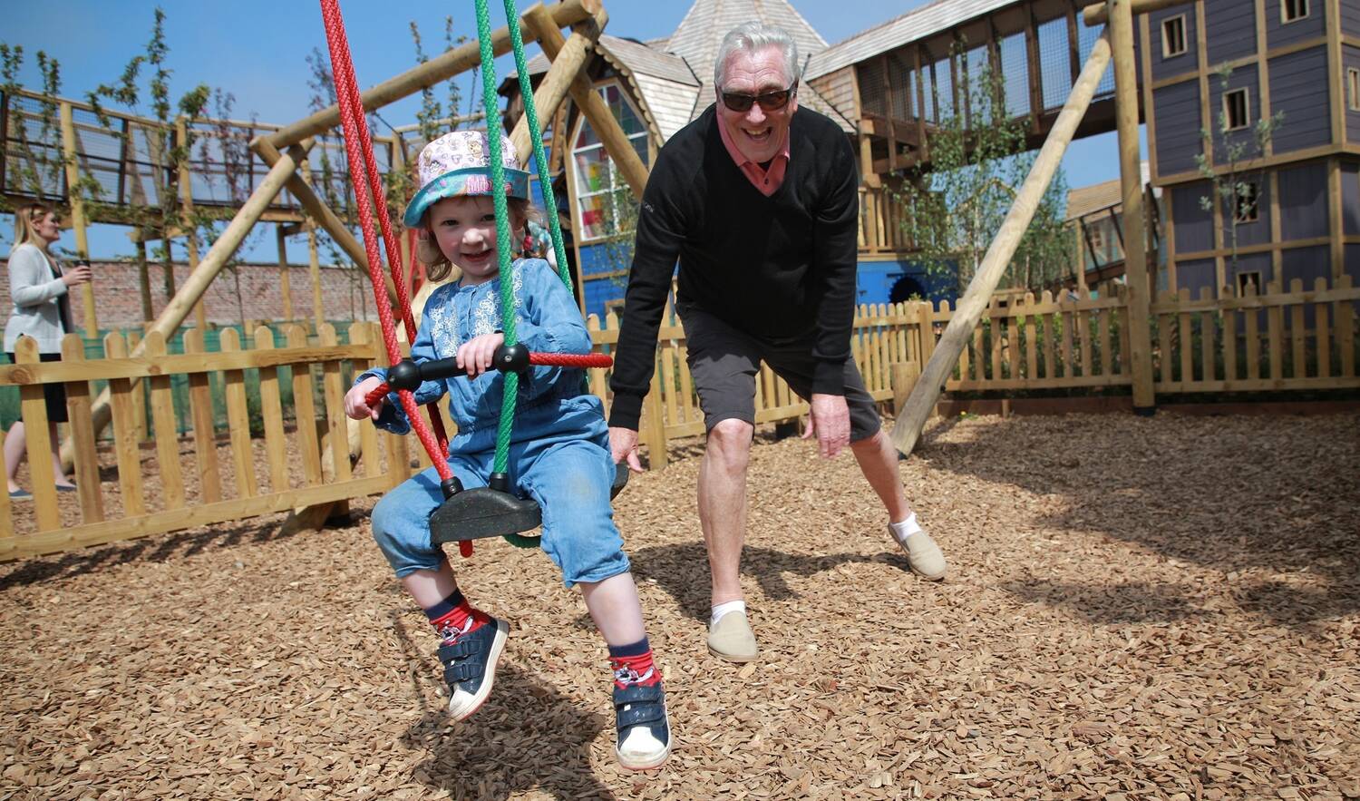 A child in a sun hat sits in a swing seat, while behind her a man with grey hair and sunglasses reaches out to push the swing for her.
