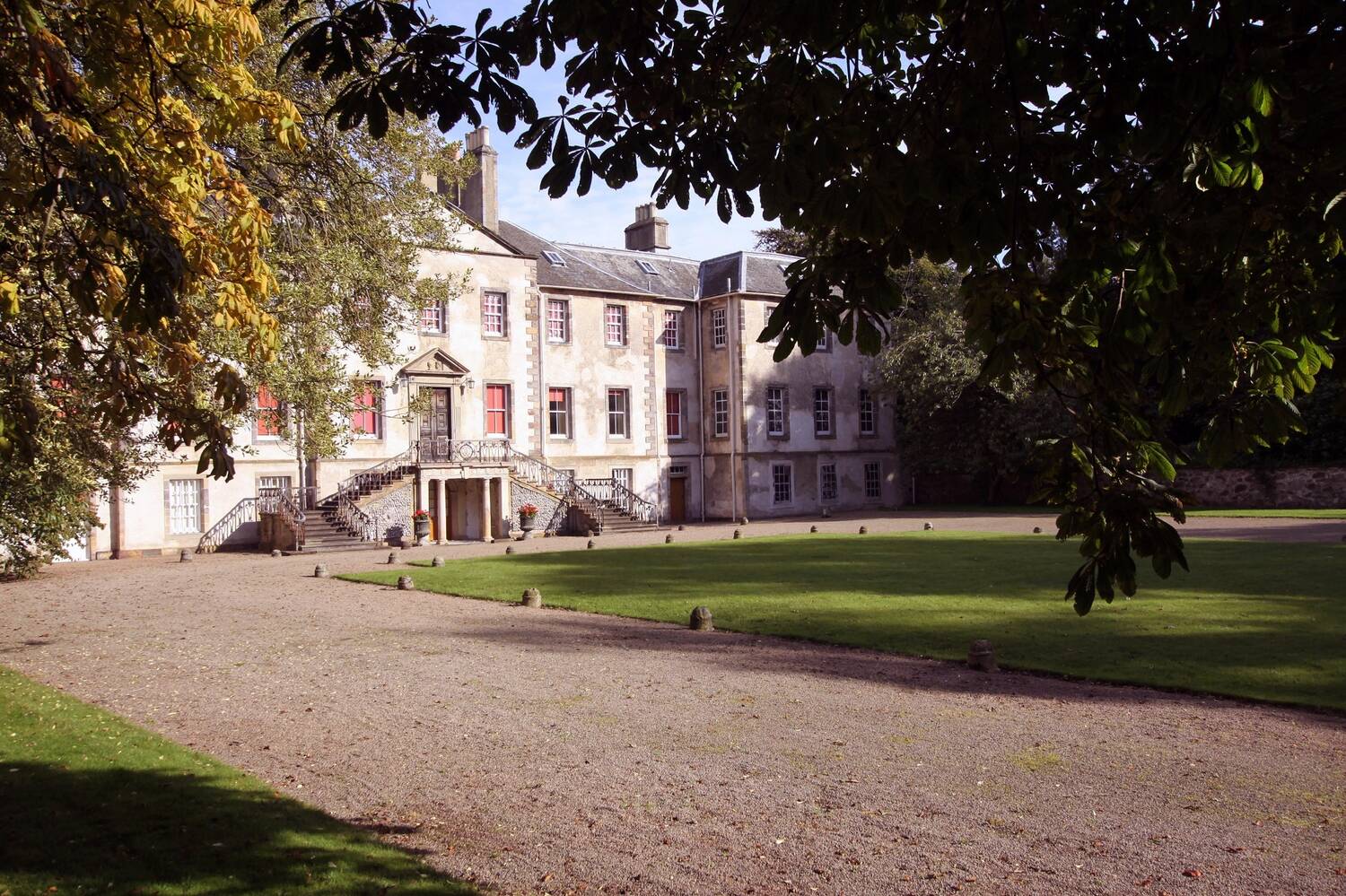 A view of the grand exterior of Newhailes House, seen from beneath leafy horse chestnut trees. A large gravel drive sweeps up to the stone entrance staircase.