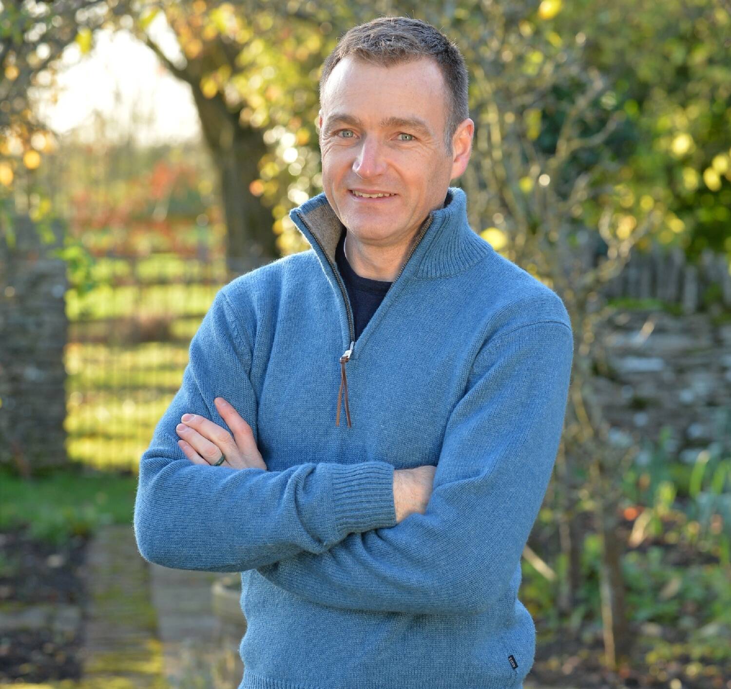 Chris Beardshaw stands in a garden, smiling at the camera with his arms folded in front of him. He wears a bright blue woolly jumper, with a half zip.