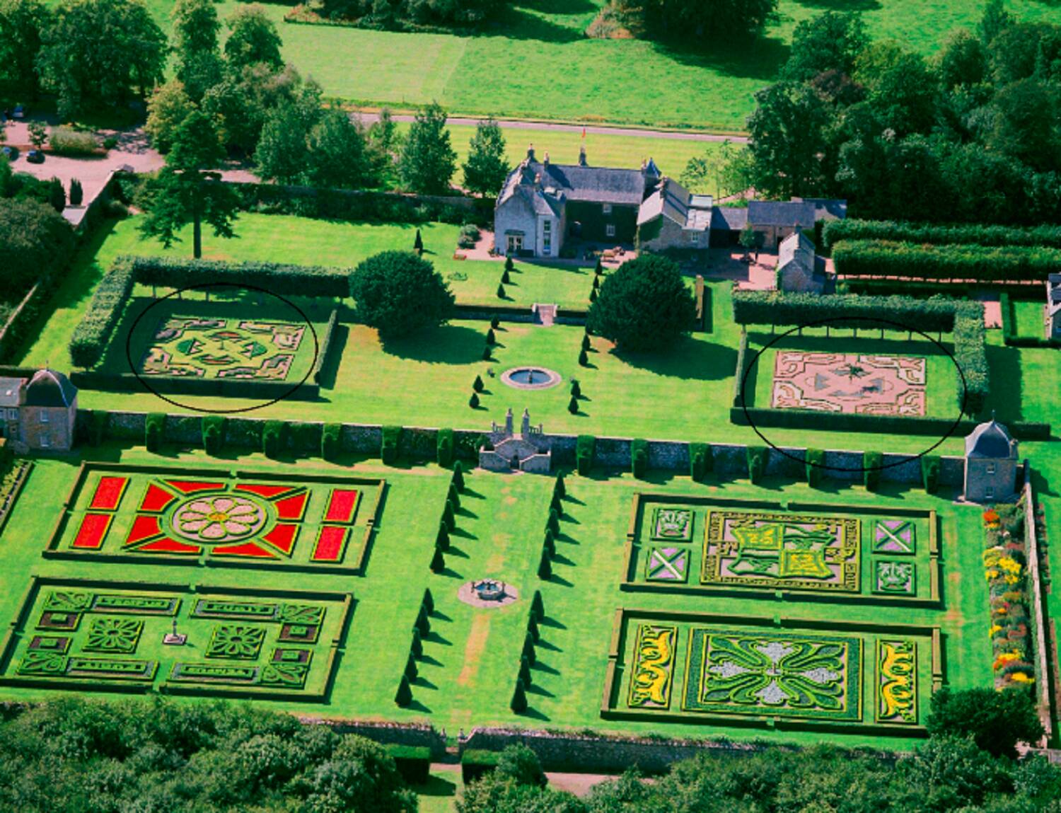 An aerial view of Pitmedden Garden, revealing the 6 distinct and colourful parterre gardens. A large stone house stands at the far end of the garden with tall trees around the perimeter.