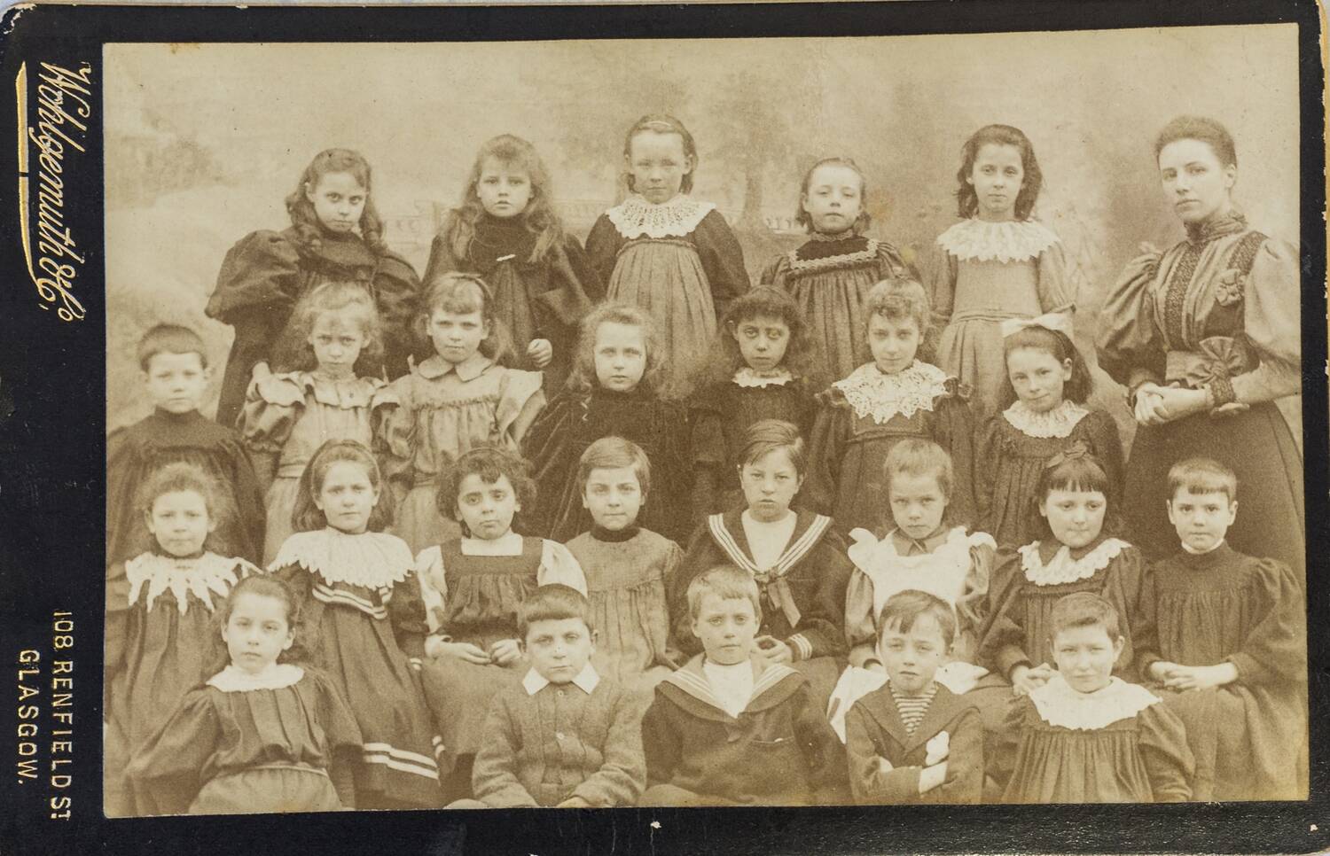 A sepia photograph of 4 rows of schoolchildren (girls and boys) with their teacher standing to the right. The children look to be aged around 7. Beside the photograph is a mount with the text: Wohlgemuth & Co, 108 Renfield St, Glasgow.