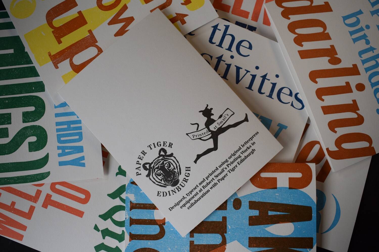 A pile of letterpress-printed cards are arranged over a flat surface. Most cards are bright and colourful, and are facing up. The card on the top of the pile is reverse-side up. It shows the logos of Paper Tiger and Robert Smail's Printing Works, as well as a couple of lines of text describing how the cards are printed.