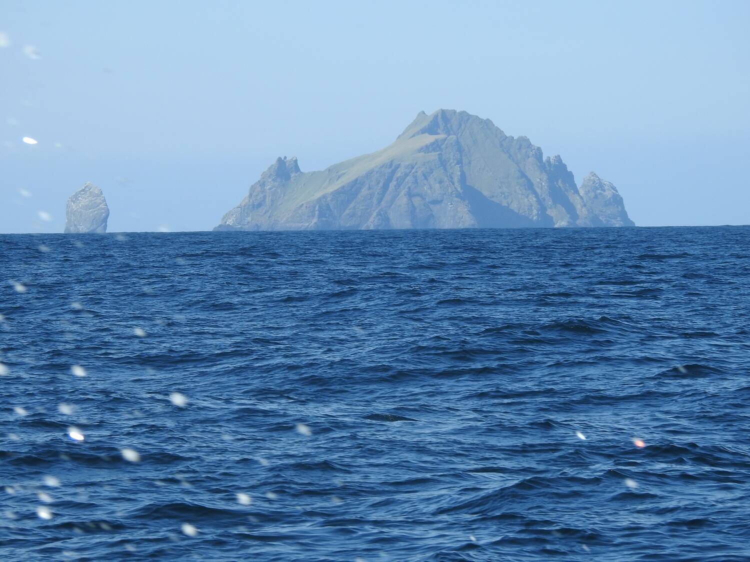 A rugged, steep-sided island rises out of the blue sea on the hazy horizon. A small island/sea stack rises just to the left of the main island. Small water droplets can be seen on the camera lens.