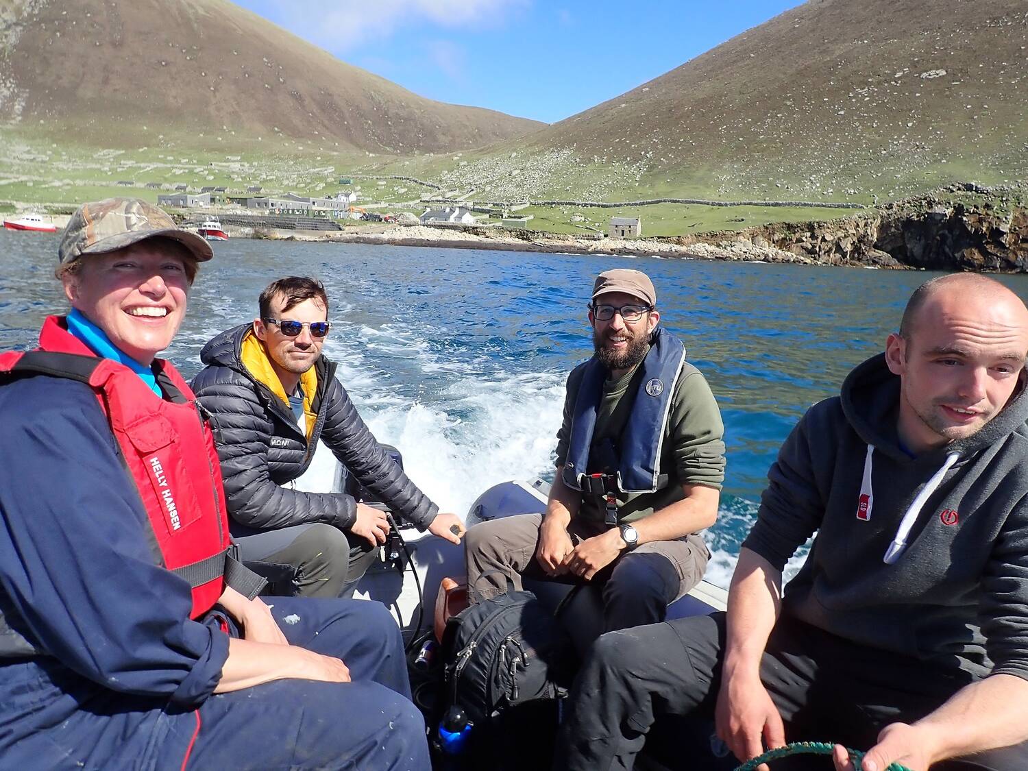 Four people sit in a RIB, heading across the sea away from an island beach. They wear life jackets and are smiling.