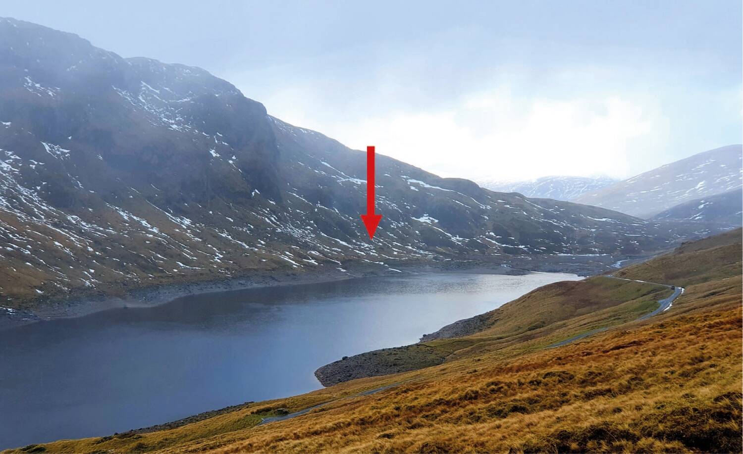 A view looking across a loch from a hillside to another steep hillside. An area on the far side of the loch has been marked by a super-imposed large red arrow.
