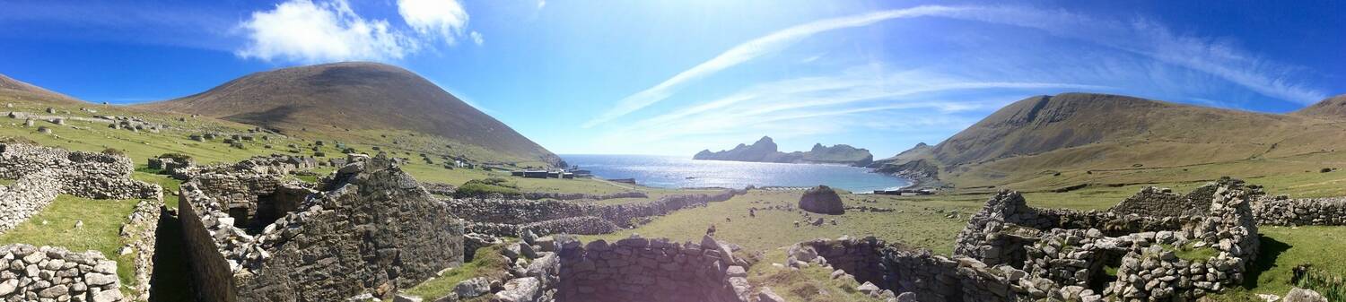A panoramic view of Village Bay in St Kilda on a sunny day with deep blue sky. In the foreground are a row of ruined stone cottages, with a large cleit standing in the field before the shore.