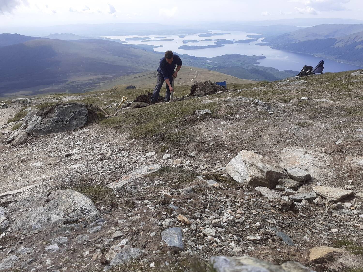 A man works on a rocky mountain summit, with Loch Lomond seen in the distance far below. He uses a sharp axe-like tool to work at the path edge.