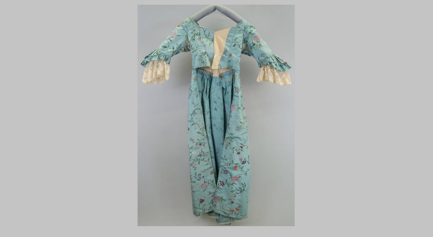 A blue silk dress with a floral pattern hangs from a padded coat hanger in front of a plain grey background. The short sleeves are finished with lace wide cuffs, and the bodice opens slightly at the front.