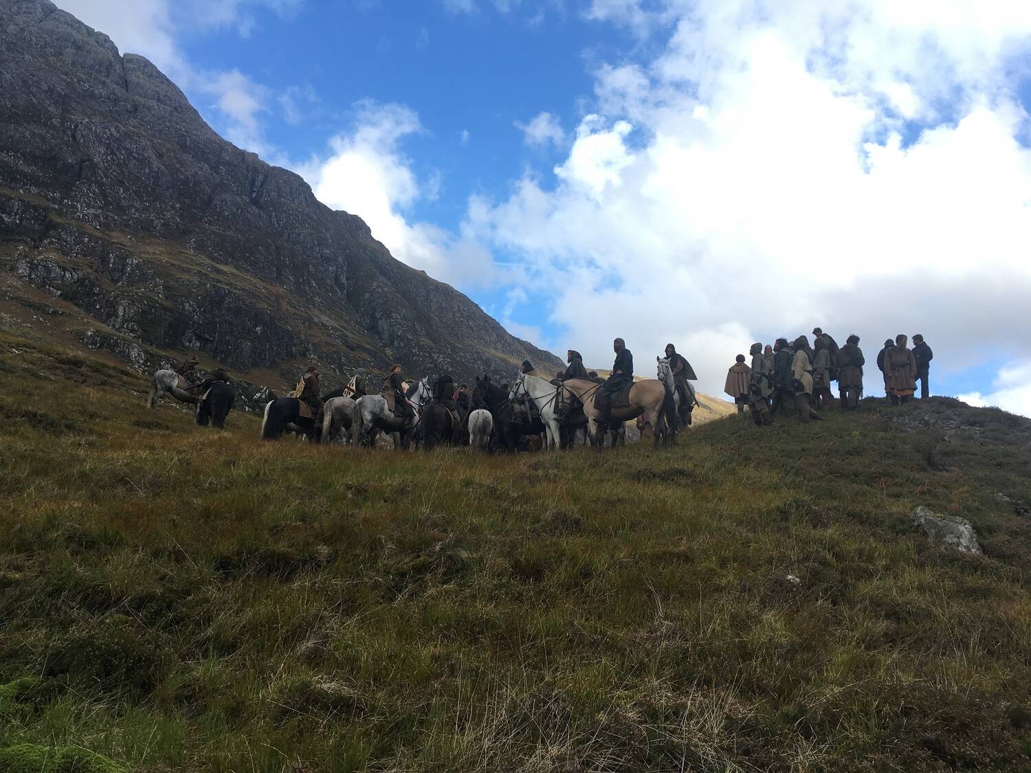 A large group of actors, many on horseback, stand on a ridge with a mountain backdrop. White clouds scatter across a blue sky.