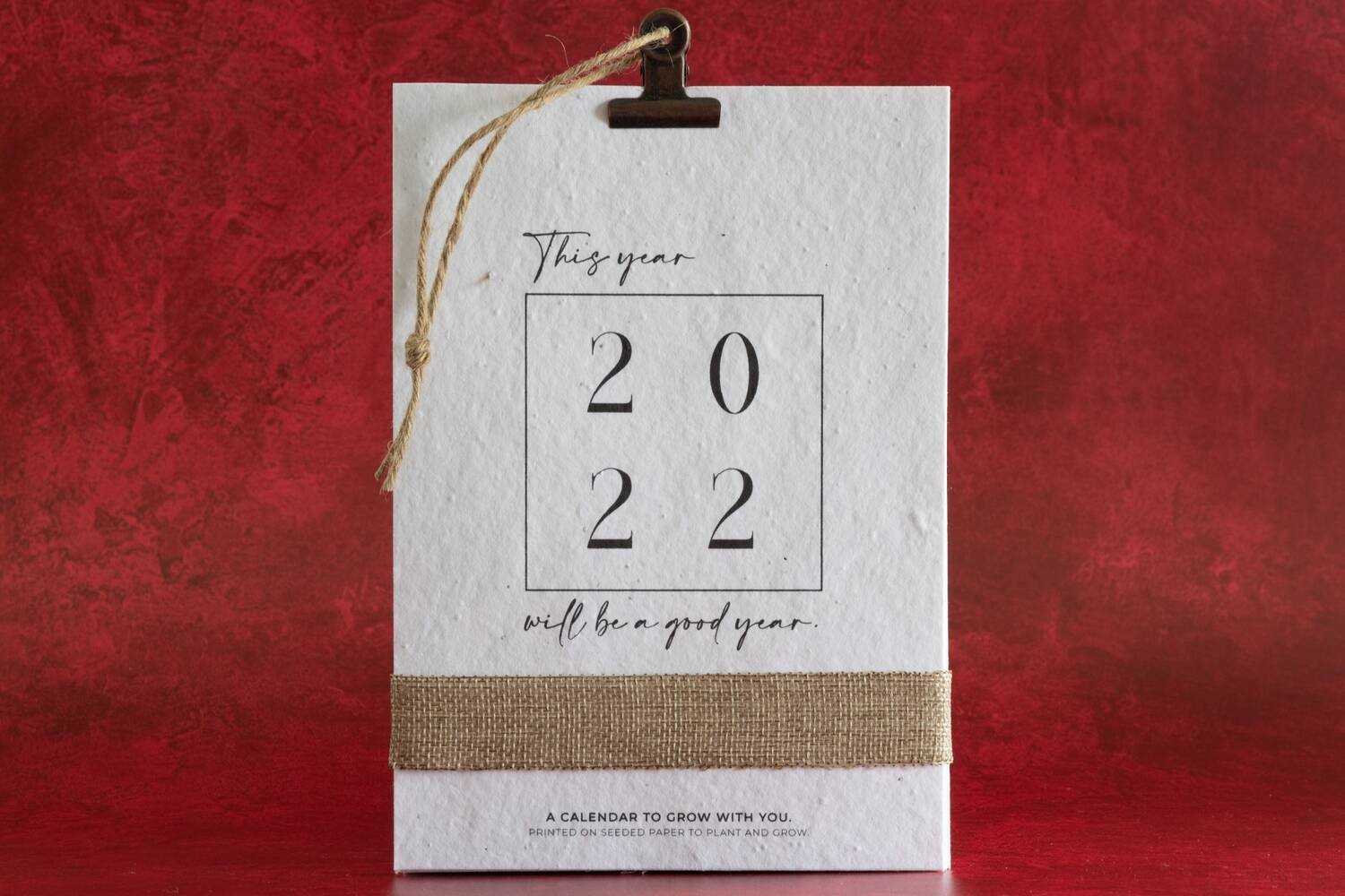 A white, square, paper calendar is held by a bulldog clip at the top and displayed against a red background. It has a hessian tie around the bottom. The front cover says: This year 2022 will be a good year. At the bottom of the page it says: A calendar to grow with you - printed on seeded paper to plant and grow.