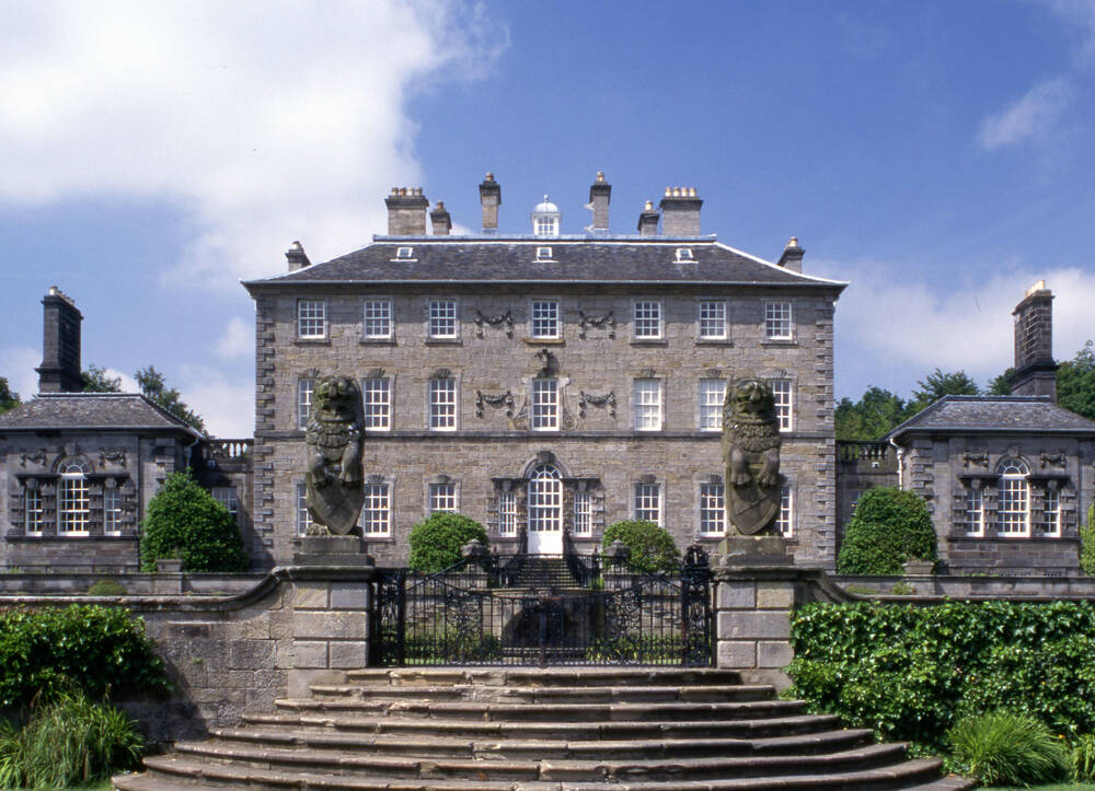 A grand 3-storey Georgian House, flanked by two single-storey pavilions. There are curving stone steps with two lion statues on pillars leading up to the house.
