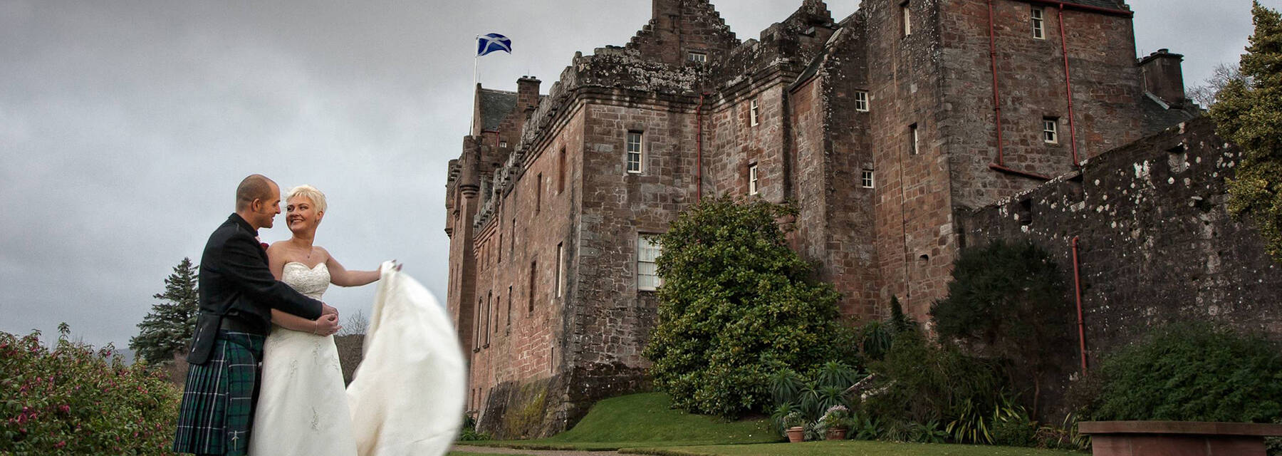 A wedding couple stand on the grassy slope outside Brodick Castle. The groom has his hands on the bride’s waist; she is holding her white dress fanned out to one side.