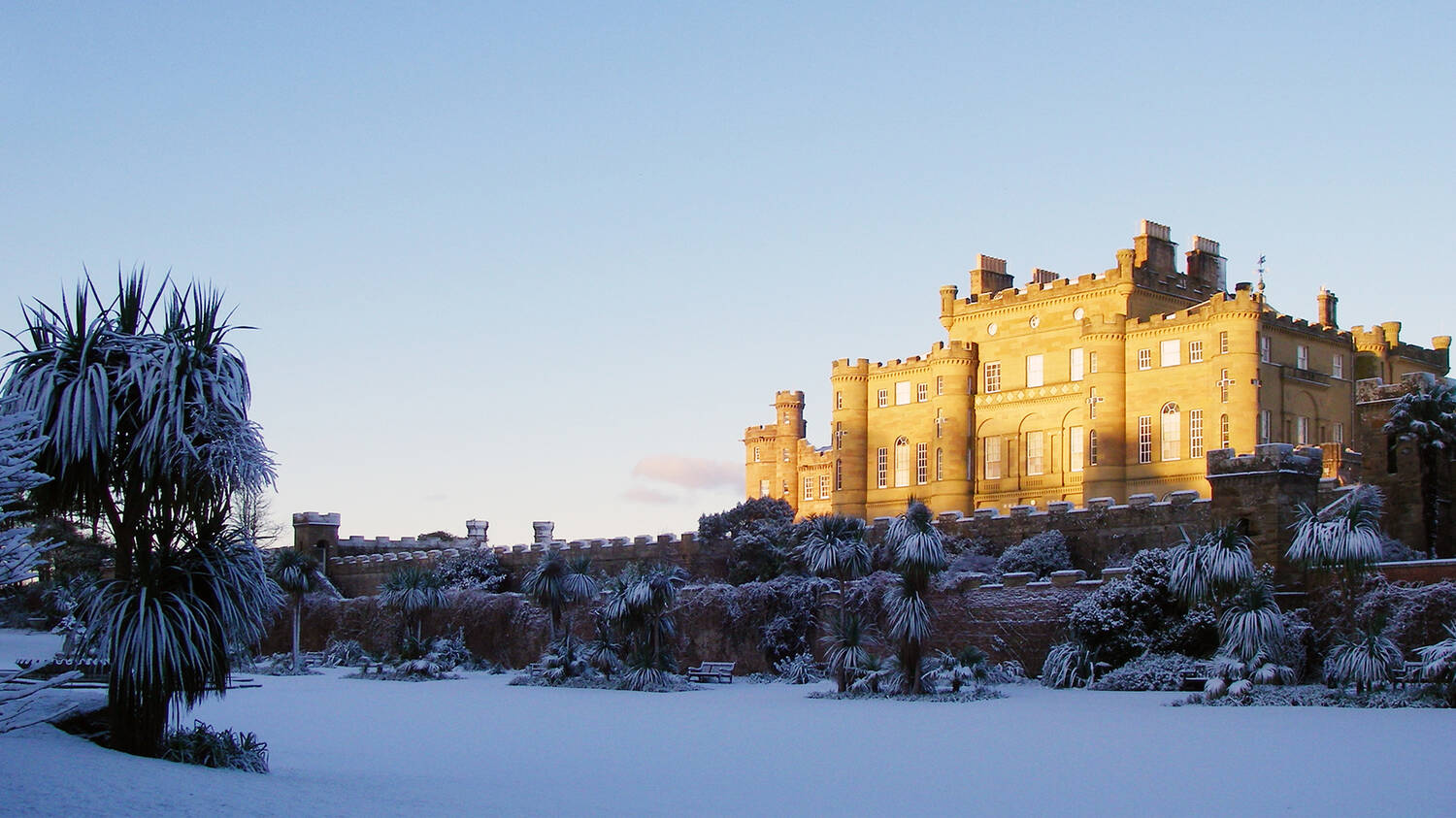 A view of Culzean Castle on a snowy day. The sun shines off the east-facing wall, making it glow orange. Thick snow covers the gardens and shrubs in the foreground, which are in the shade.