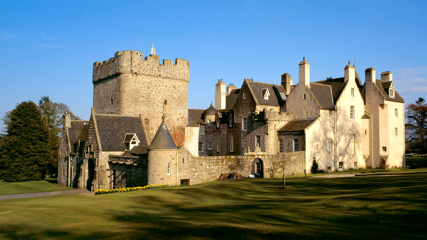 A view of Drum Castle on a sunny day, with bright blue sky behind. The ancient, square stone tower is to the left, with the Jacobean section to the right. Tall trees can be seen behind the castle.